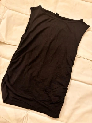 Svart ärmlös linnetopp - En enkel och stilren svart linnetopp med rund halsringning och ärmlös design. Toppen har en lätt rynkad detalj på ena sidan som ger en snygg twist. Perfekt att matcha med jeans eller kjol för en clean look.