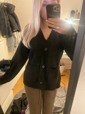 Svart kofta - En svart kofta med v-ringning och stora knappar framtill. Koftan har två fickor och ribbade muddar vid ärmslut och nederkant. 