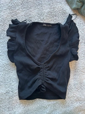 Svart ribbad topp från Zara - Säljer en svart ribbad topp från Zara med v-ringning, rynkad snörning framtill och volangdetaljer på de korta ärmarna. Perfekt för dig som gillar trendiga och feminina detaljer. Superfin att styla till både jeans och kjol!