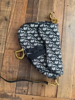 Dior väska - Ikonisk Dior Saddle Bag i svart och vitt canvasmaterial med monogrammönster. Väskan har guldfärgade metalldetaljer, bland annat det klassiska D-spännet, och svarta läderdetaljer. Unik sadelformad design och justerbar axelrem.