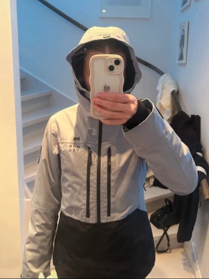 Skidjacka från MONTEC - Snygg skidjacka från MONTEC i grå/blå och svart med huva och dragkedjor framtill. Jackan har två vertikala bröstfickor med svarta dragkedjor.