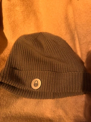 Sail Racing Race Beanie – Grå stickad mössa i toppskick - Säljer en brun ribbstickad mössa med uppvikt kant och en rund patch framtill med ett stilrent tryck. Mössan är i mjukt, stretchigt material och passar perfekt för kyliga dagar. Enkel och clean design som funkar till det mesta.