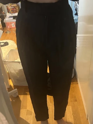 Svarta kostymbyxor med hög midja - Säljer ett par svarta kostymbyxor med hög midja och avsmalnande ben. Byxorna har knapp och dragkedja framtill samt diskreta fickor i sidorna. Perfekta för en clean och stilren look. 