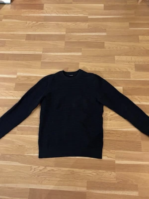 Mörkblå tröja J.Lindeberg - Snygg mörkblå stickad tröja från J.Lindeberg med diskret rutstruktur. Tröjan har rund halsringning och ribbade muddar vid ärmslut och nederkant. Perfekt för dig som gillar stilrena och klassiska plagg.