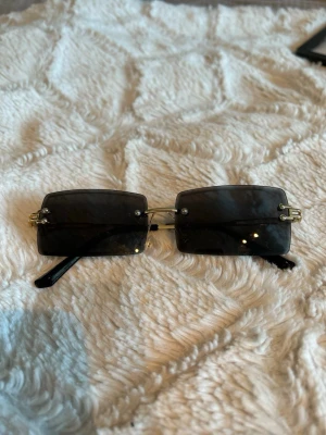 Svarta rektangulära solglasögon - Stilrena solglasögon med rektangulära svarta glas och tunna guldfärgade bågar. Glasen är mörka och bågarna har minimalistisk design utan synlig ram runt glasen. Perfekta för en trendig look.