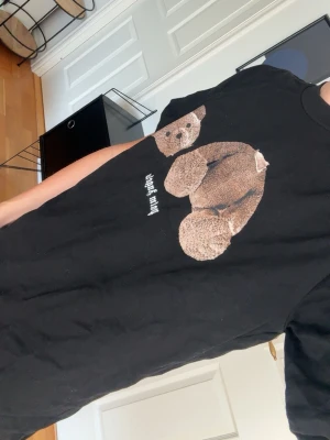 Palm Angels svart t-shirt med björn - Svart t-shirt från Palm Angels med ikoniskt björntryck på framsidan och stor vit logga på ryggen. Klassisk passform och rund hals. Perfekt statement-plagg för dig som gillar streetwear och coola prints.