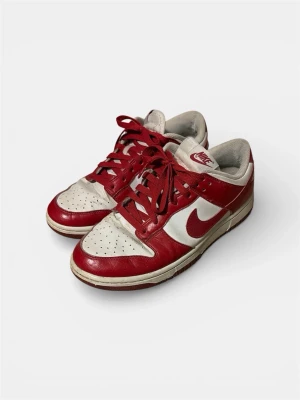 Nike Dunk Low röd/vit sneakers - Nike Dunk Low sneakers i rött och vitt läder med klassisk swoosh på sidan. Skorna har perforerad tåbox, röda skosnören och vit mellansula. Ikonisk retrostil som passar perfekt till streetwear och sneakersamlingen. Storlek: 40.5