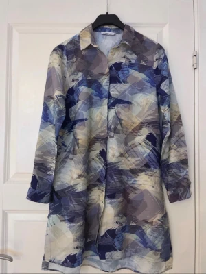 Blåmönstrad skjortklänning - Unik skjortklänning med abstrakt blått, grått och beige mönster. Klänningen har krage, knappar framtill och långa ärmar. Perfekt för dig som gillar konstnärliga prints och en avslappnad men snygg stil.