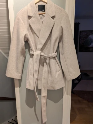 Ljusbeige kappa med knytskärp - Stilren ljusbeige kappa från H&M med bred krage och knytskärp i midjan. Kappan har rak passform, långa ärmar och är tillverkad i ett mjukt tyg som ger en clean look. Perfekt för dig som gillar minimalistisk stil.