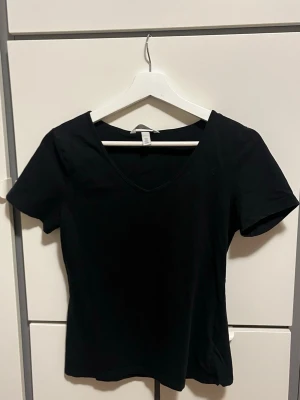 Svart v-ringad t-shirt från H&M - En stilren svart t-shirt från H&M med v-ringning och korta ärmar. Modellen har en klassisk passform och är tillverkad i bomull, perfekt för en enkel och clean look.