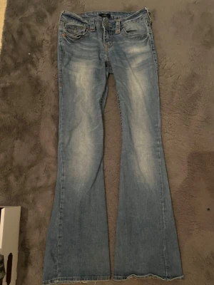 Blå bootcut jeans från True Religion - Säljer ett par ljusblå bootcut jeans från True Religion med slitningar och snygga detaljer på bakfickorna. Jeansen har klassisk femficksmodell, låg midja och kontrastsömmar. Perfekta för dig som gillar en avslappnad men trendig look. Dragkjedjan går ner för mig! Tror det är för att dem är för små! 