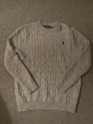  Stickad  Polo Ralph Lauren - Kabelstickad beige tröja från Polo Ralph Lauren med rund halsringning och mörkblå broderad logga på bröstet. Tröjan har långa ärmar och ribbade muddar vid ärmslut och nederkant. Perfekt för lager på lager och klassisk stil.
