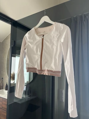 Vit cropped ziptröja från Aim'n - Snygg vit långärmad ziptröja från Aim'n med croppad passform och beige resår med logga i midjan. Tröjan har rund halsringning och dragkedja framtill. 