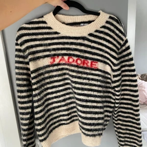 Randig stickad tröja med J'ADORE - Säljer en svartvit randig stickad tröja med texten 'J'ADORE' i rött framtill. Tröjan har rund halsringning, långa ärmar och ribbade muddar. Perfekt för dig som gillar en trendig och mysig look.