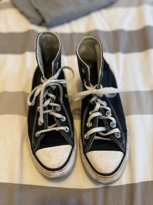 Svarta Converse Chuck Taylor Highs - Klassiska svarta Converse Chuck Taylor All Star med högt skaft, vita gummisulor och tåhättor samt vita snören. Skorna har den ikoniska loggan på sidan och är tillverkade i slitstark canvas. Perfekta för en avslappnad och cool stil.