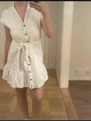 Vit kort klänning med knappar - Söt vit kortklänning med broderade detaljer, knappar framtill och knytband i midjan. Klänningen har v-ringning, volangkant nertill och är kortärmad. Perfekt för varma dagar och ger en fräsch sommarkänsla.