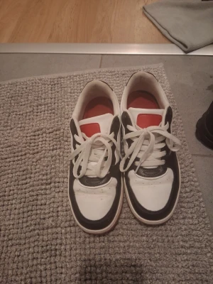 Svarta och vita sneakers med röd sula - Säljer ett par stilrena sneakers i svart och vitt med röda detaljer och röd sula. Skorna har klassisk låg profil, vita snören och paneler i syntetmaterial. Perfekta för dig som gillar en sportig look med kontrastfärger.