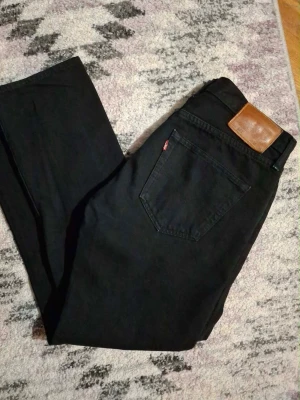 Svarta Levi's 501 jeans - Klassiska svarta Levi's 501 jeans med rak passform och fem fickor. Jeansen har den ikoniska läderpatchen bak i midjan och röd Levi's-tag på bakfickan. Tillverkade i slitstark denim, perfekta för en tidlös och avslappnad look.