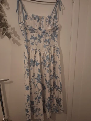 Vit och blå blommig långklänning - Romantisk långklänning med blått blommönster på vit botten. Klänningen har smala axelband med knyt, volang upptill och smockad rygg för skön passform. Perfekt för sommarens alla tillfällen.