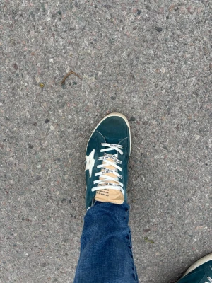 Gröna Golden Goose sneakers med stjärna - Snygga gröna sneakers från Golden Goose med vit stjärndetalj på sidan och vita skosnören med text. Skon har en beige plös och klassisk låg silhuett. Perfekt för dig som gillar streetstyle och vill ha något unikt på fötterna.