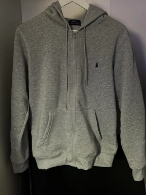 Grå zip-hoodie från Polo Ralph Lauren - Säljer en grå hoodie med dragkedja från Polo Ralph Lauren. Tröjan har huva med snören, två fickor fram och den klassiska logon broderad på bröstet. Perfekt för chill dagar och enkel att matcha med jeans eller joggers.