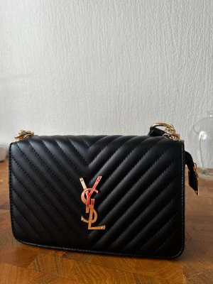 Svart YSL axelväska med guldkedja - Snygg fejk svart axelväska från YSL i quiltat läder med guldfärgade detaljer och kedjerem. Väskan har det klassiska YSL-emblemet framtill och en stilren, rektangulär form. Perfekt för dig som vill ha en lyxig och trendig accessoar.