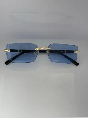 Rektangulära blå solglasögon guld - Snygga solglasögon med rektangulära, blåtonade glas och tunna guldfärgade detaljer. Glasögonen har minimalistisk design utan synlig båge runt glasen och svarta skalmar. Perfekt accessoar för en trendig look.