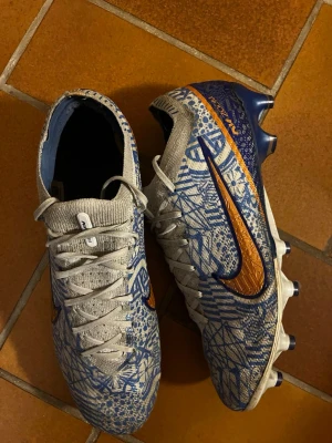 Nike Mercurial fotbollsskor blå/orange - Säljer ett par Nike Mercurial fotbollsskor med grå bas, blå grafiskt mönster och orange detaljer. Skorna har klassisk snörning, låg profil och dobbar för spel på gräs. Swoosh-loggan i orange och blått på sidan ger en riktigt snygg look. Perfekta för dig som vill sticka ut på planen.