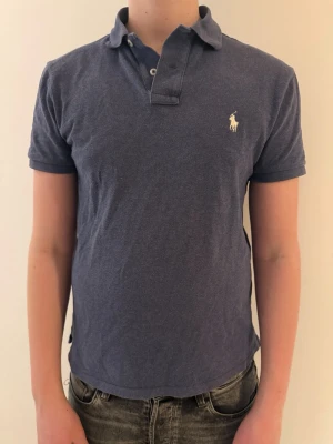 Blå pikétröja från Ralph Lauren - Klassisk blå pikétröja från Ralph Lauren med korta ärmar och vit broderad logga på bröstet. Tröjan har krage och knäppning framtill. Perfekt för en avslappnad och stilren look. Det finns ett ihop sytt hål men som inte längre syns. Storlek S