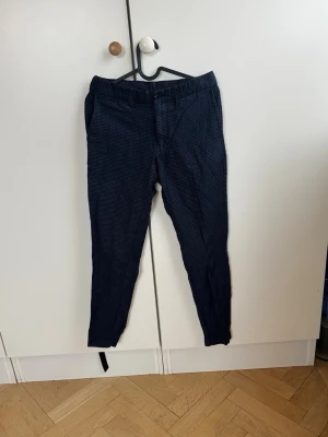 Marinblå rutiga kostymbyxor J.Lindeberg  - Snygga marinblå kostymbyxor med diskret rutmönster och smal passform. Byxorna har resår i midjan, sidfickor fram och två bakfickor med knapp. Perfekta för en stilren look och passar till skjorta eller t-shirt. Nypris 1600