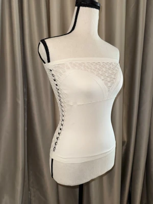 Vit bandeau tubtopp från Guess - Säljer en vit bandeau tubtopp från Guess med snygga spetsdetaljer framtill och längs bak. Toppen är axelbandslös och har en tajt passform som framhäver figuren. 