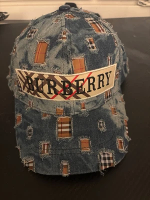 Burberry denimkeps med patchwork - Säljer en unik Burberry keps i blå denim med slitna detaljer och patchwork i klassiskt rutmönster. Stor Burberry-logga framtill och justerbart band bak i svart läder med präglad logotyp. Perfekt för dig som vill sticka ut med en cool accessoar. Pris kan diskuteras 