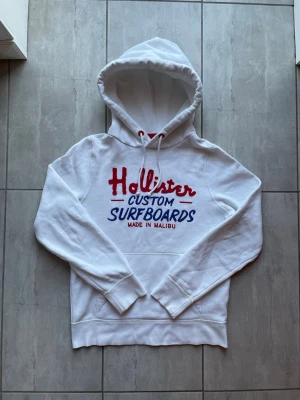 Vit hoodie från Hollister med tryck - Vit hoodie från Hollister med rött och blått broderat tryck på bröstet där det står 'Hollister Custom Surfboards Made in Malibu'. Klassisk känguruficka och dragsko i huvan. Perfekt för en avslappnad och cool stil.