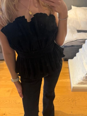 Svart axelbandslös volangtop - Trendig svart axelbandslös topp med volangdetaljer och markerad midja. Toppen har en unik struktur och ger en snygg siluett, perfekt för dig som vill sticka ut. Passar till både jeans och kjol för en cool look.