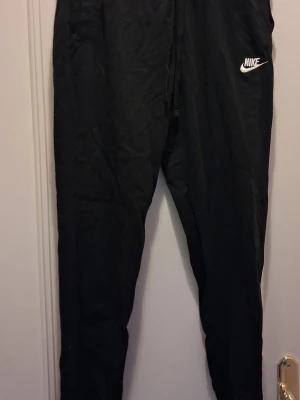 Svarta mjukisbyxor från Nike - Svarta mjukisbyxor från Nike med dragsko i midjan och smal passform. Byxorna har en liten vit Nike-logga på vänster lår och är tillverkade i ett mjukt och bekvämt material. Perfekta för chill eller träning.