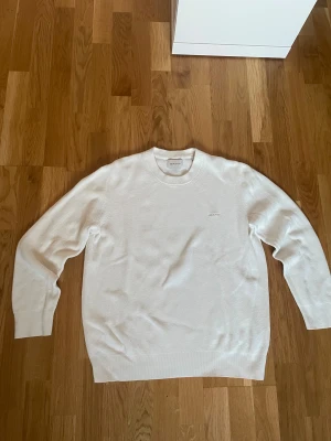 Vit stickad tröja från GANT - Vit stickad tröja från GANT i storlek M. Tröjan har rund halsringning, diskret GANT-logga på bröstet och är tillverkad i mjuk bomull. Klassisk och clean look med långärmade ribbade muddar och strukturstickat material.