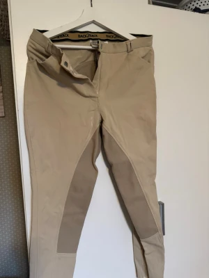 Beige ridbyxor från Back2Back - Snygga beige ridbyxor från Back2Back med förstärkta partier på insidan av benen. Byxorna har en klassisk passform, bälteshällor och dragkedja med knapp. Perfekta för dig som gillar hästar och vill ha bekväma byxor i stallet.