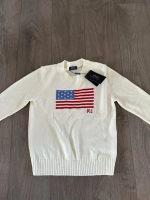 Vit stickad tröja Polo Ralph Lauren - Vit stickad tröja från Polo Ralph Lauren med amerikansk flagga i blått, rött och vitt på bröstet samt RL-broderi. Tröjan har rund halsringning och ribbstickade muddar vid ärmslut och nederkant. Klassisk och stilren design.