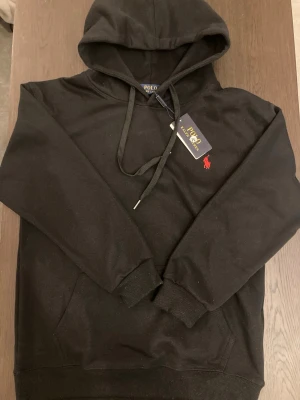 Svart hoodie från Polo Ralph Lauren - Svart hoodie från Polo Ralph Lauren med klassisk huva och dragsnören. Broderad röd logga på bröstet och ribbade muddar vid ärmslut och nederkant. Mjuk bomullskänsla och känguruficka framtill. Perfekt för en chill och stilren look. Storlek M men tycker själv den är lite liten i storleken då jag är ganska smal. Så du får tänka dig att det är en mindre medium.