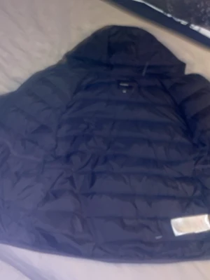 Svart pufferjacka från Lyle & Scott - Snygg svart pufferjacka från Lyle & Scott Junior. Jackan har en klassisk quiltad design med hög krage och är perfekt för kalla vinterdagar. Tillverkad i ett glansigt material som ger en modern look. Passar dig som vill ha både stil och värme. De ända som är fel med den är att den har en liten bit grön färg på en av armarna men det är knappt synligt 