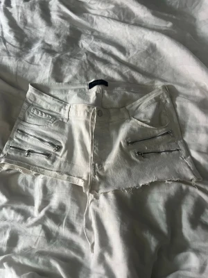 Vita denimshorts med dragkedjor - Det är en stor storlek men det finns snören som man knyter, så den passar alla.Säljer ett par vita denimshorts med rå kant och fyra silverfärgade dragkedjor framtill. Shortsen har hög midja, klassiska fickor både fram och bak samt en cool, edgy look. Perfekta för sommaren och festivaler!