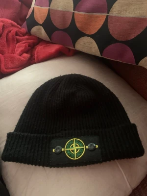  Stone Island mössa svart  - Säljer en svart ribbstickad mössa från Stone Island med den klassiska logotypen framtill på en patch. Mössan har uppvikt kant och är tillverkad i mjukt material som sitter skönt på huvudet. Perfekt accessoar för att höja din streetwear-stil.