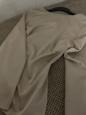 Beige blus med knytdetalj - Stilren beige blus med lång ärm och snygg knytdetalj i sidan. Blusen har en mjuk, något glansig yta och rund halsringning. Perfekt för dig som gillar en enkel men trendig look.