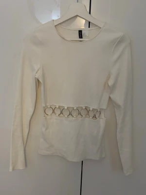 Vit långärmad topp med ringdetaljer - Vit långärmad topp från H&M med guldfärgade ringdetaljer i midjan. Toppen är ribbad och har en figurnära passform, säljs på grund av knapp användning, endast använd en gång💕