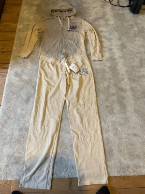 Beige Lexington loungewear jumpsuit - Mysig beige jumpsuit från Lexington med huva, dragkedja framtill och långa ärmar. Byxdelen har resår och snörning i midjan samt tryckt Lexington-logga på bröstet och benet. Perfekt för chill dagar hemma. Huvtröjan är i storlek L och byxorna i storlek XL. Helt nytt mjukissett. Nypris 2200kr