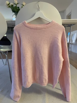 Ljusrosa stickad tröja - Mysig ljusrosa stickad tröja med rund halsringning och långa ärmar. Tröjan har en enkel och stilren design med ribbstickade muddar vid hals, ärmslut och nederkant. Perfekt för att hålla värmen under kyliga dagar.