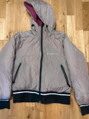 Beige vindjacka från Sailracing med huva - Snygg beige vindjacka från Sail Racing med svart dragkedja, två snedställda fickor  , Racing-logga på bröstet. Jacka från sail  Racing har huva med dragsko och svart/vita muddar vid ärmar och sop Racing nederkant. Insidan av huvan är vinröd.