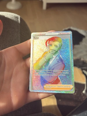 Pokemon Rose Trainer Supporter-kort - Säljer ett holografiskt Rose Trainer Supporter-kort från Pokemon Trading Card Game. Kortet har en glittrande yta med färgglada effekter och motiv av karaktären Rose. Perfekt för samlare eller dig som vill boosta din lek med en unik supporter.