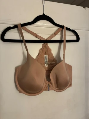 Beige bh med spetsdetal stativ Lilja - Snygg beige bh med vadder confidently kupor och bygel från Lilja, PSA easy frontknäppning. Axelbanden är justerbara och korsade i ryggen som ger extra support. Baksidan har fina spetsdetaljer som gör bh:n både stilren och feminin. 