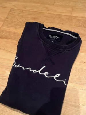 Svart sweatshirt från Bondelid - Cool svart sweatshirt från Bondelid med vit broderad logotyp på bröstet. Tröjan har rund halsringning, ribbade muddar och är långärmad. Perfekt för en avslappnad och trendig look.